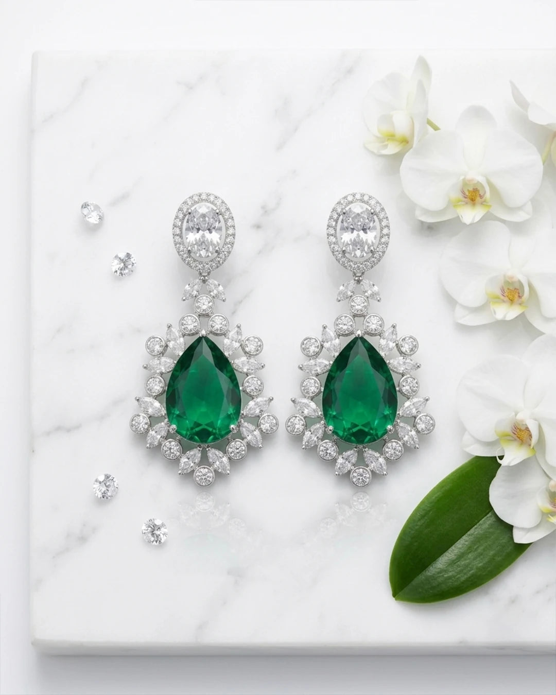Emerald green teardrop earrings with diamond-like stones on a marble surface with white flowers.