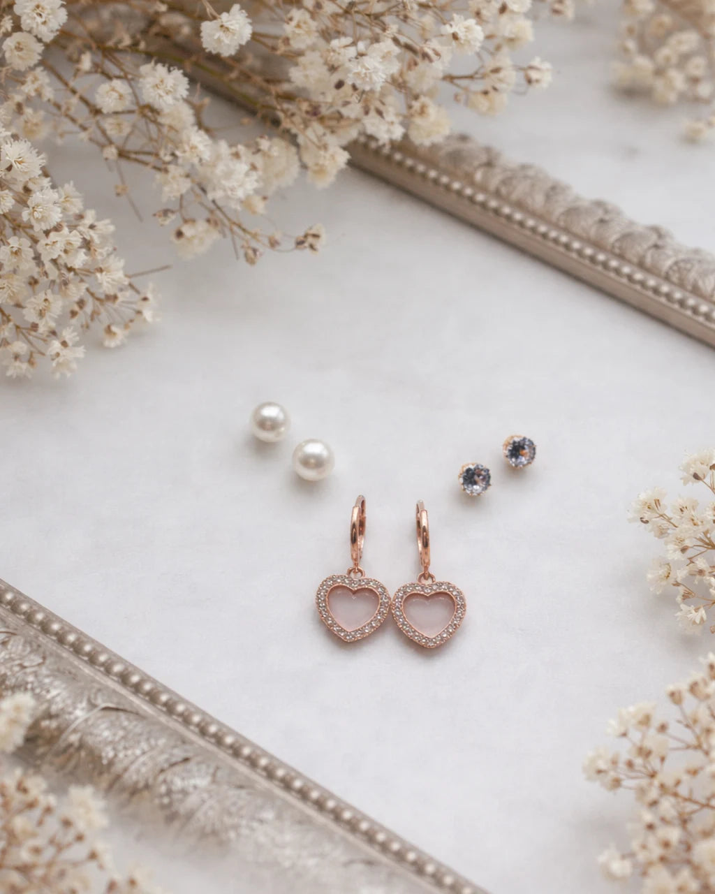 Rose gold heart-shaped earrings on a white surface with dried flowers and a decorative frame.