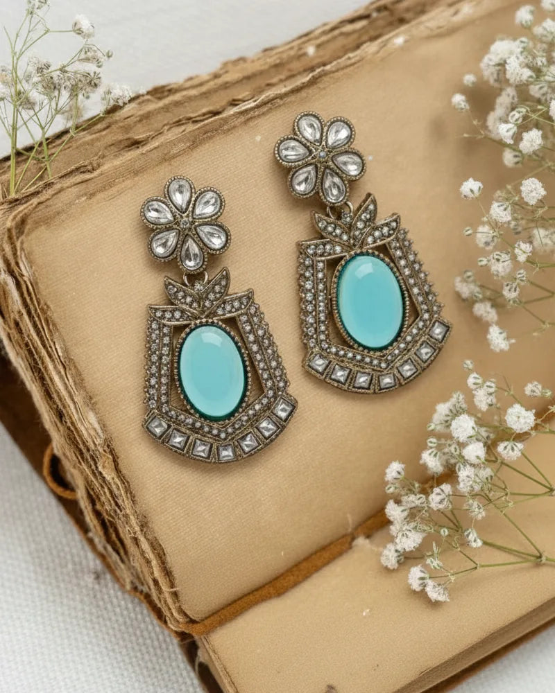 Pair of ornate earrings with turquoise stones on a textured surface with small white flowers.