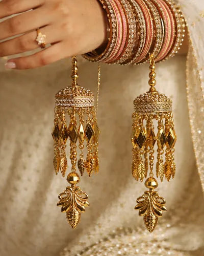 Gold earrings with intricate designs held by a hand wearing multiple bangles against a textured gold background.