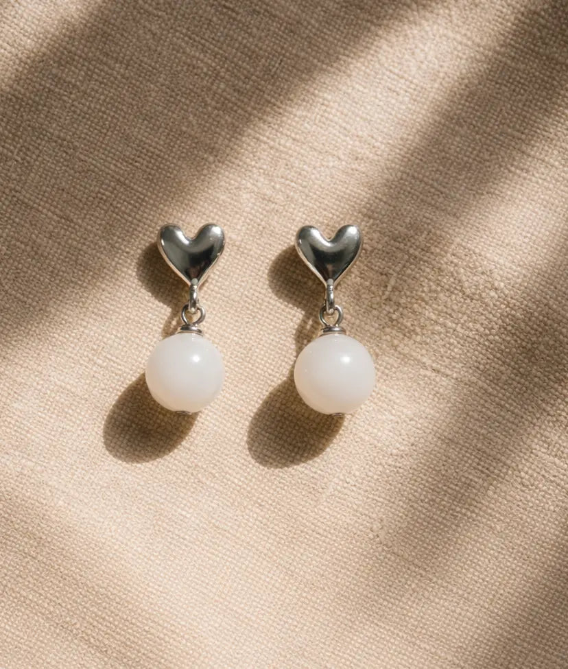 Pair of heart-shaped earrings with white spherical beads on a beige fabric background