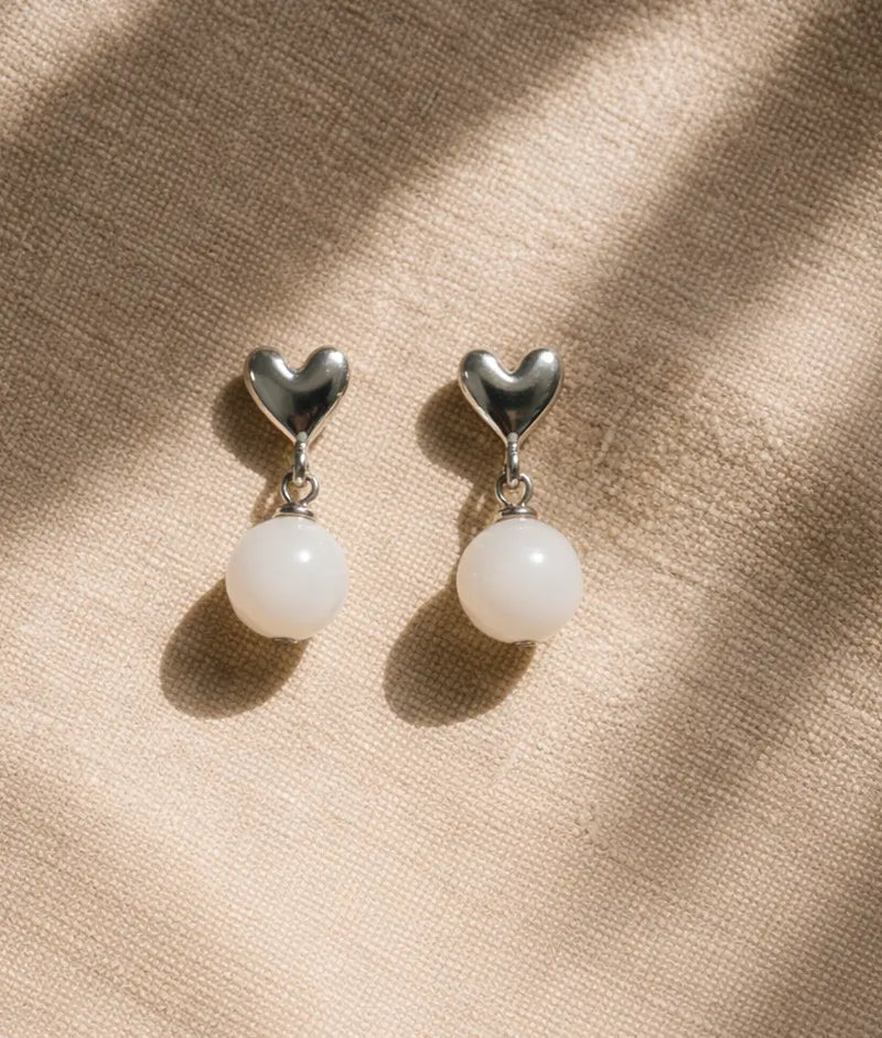 Pair of heart-shaped earrings with white spherical beads on a beige fabric background