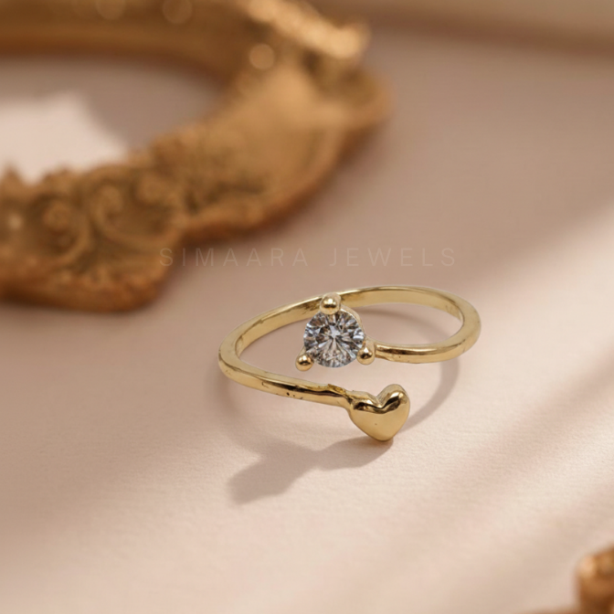Gold ring with a heart design and gemstone on a beige surface, branded 'Simaara Jewels'.