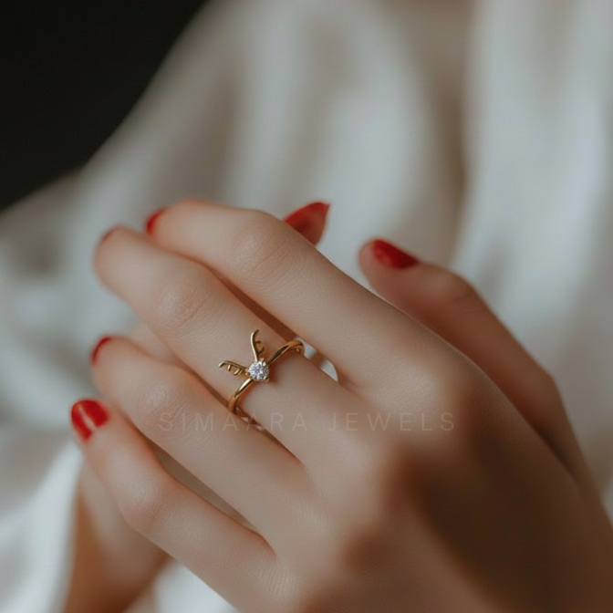 Gold ring with a diamond on a hand with red nail polish against a neutral background, featuring the brand 'Simaara Jewels'.