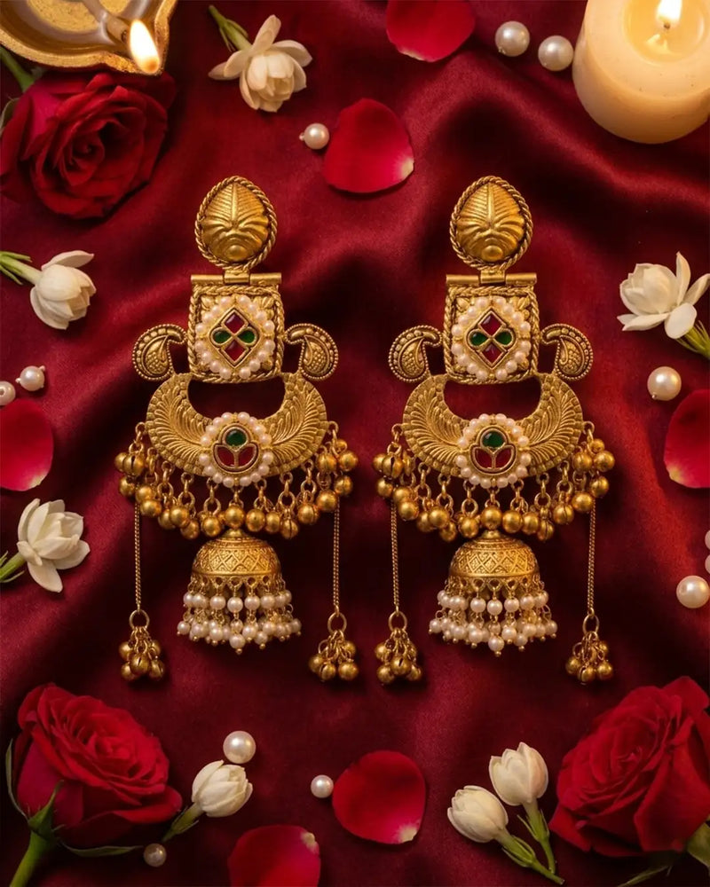 Gold earrings with red and green gemstones on a red fabric background with flowers and candles.