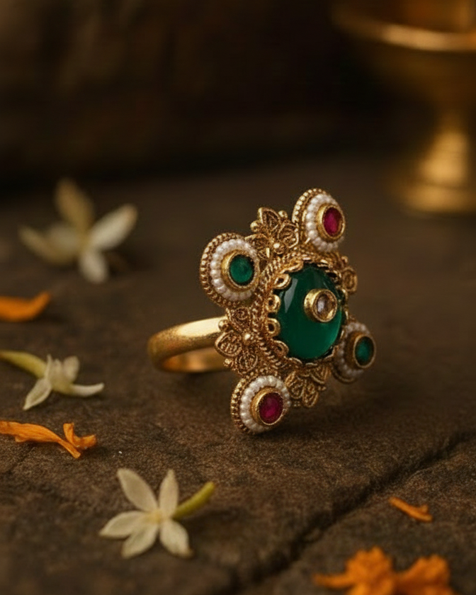 Decorative ring with gold, green, and red stones on a dark surface with flowers.